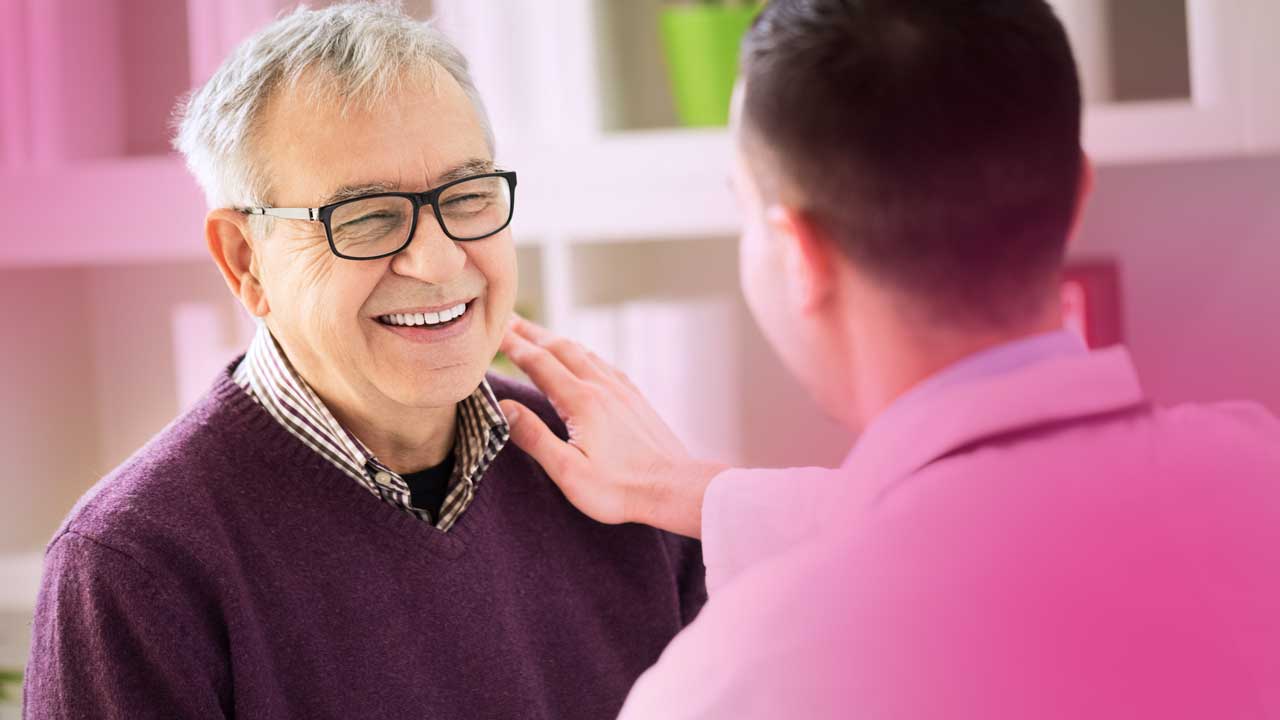 Smiling elderly man wearing glasses and a purple sweater with a healthcare professional touching his shoulder.
