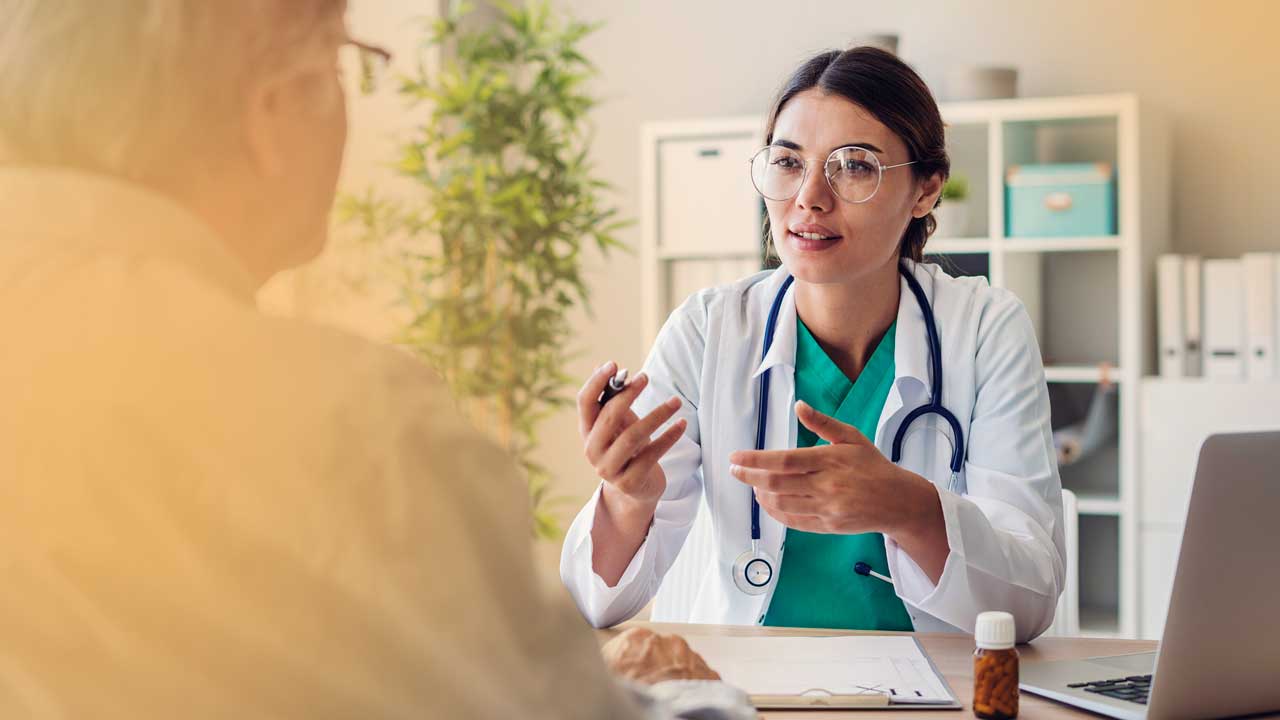 Female doctor wearing glasses and a stethoscope talking to an elderly patient in a medical office.